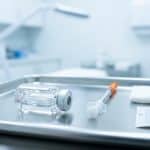 Steril tepside kapaklı ince enjektör yanında etiketsiz botulinum toksin flakonu, modern dermatoloji odası arka planı